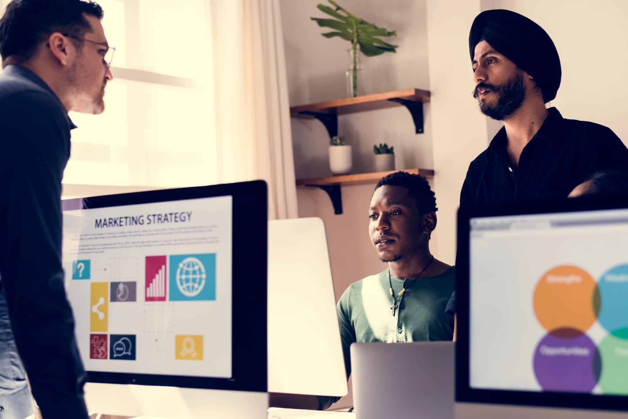 Team collaborating on a marketing strategy presentation in a modern office, with analytics displayed on desktop screens.