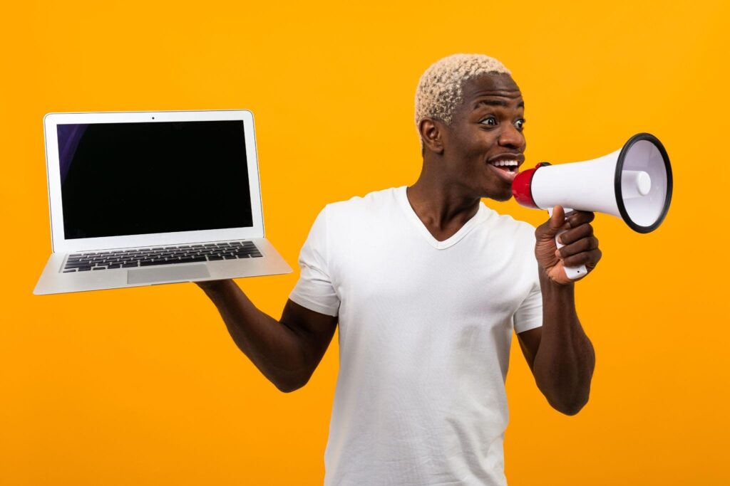 Smiling man holding a laptop and speaking into a megaphone against a bright orange background, representing an online announcement or digital marketing promotion.